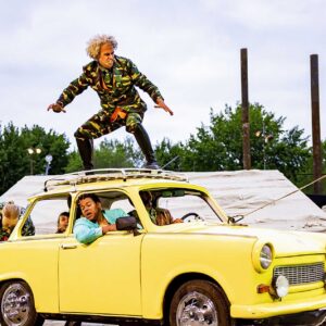 Een gele auto waar mensen in zitten, een persoon hangt met zijn ellenboog uit het raam. Bovenop staar een man in een camouflagepak.