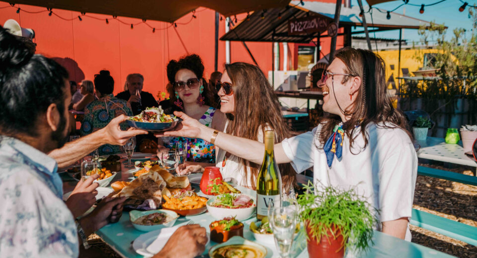 FUJA8557 (1) Mensen zitten buiten aan een tafel en genieten van een maaltijd, iemand geeft een kom eten aan een ander.