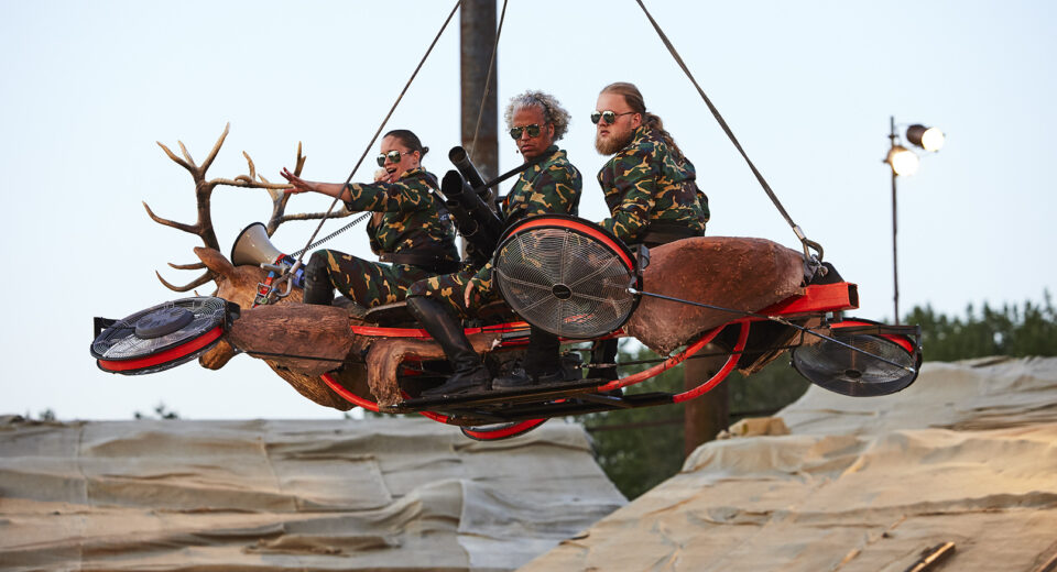 20230707_POPPY_©_DAISY_KOMEN_PHOTOGRAPHY_23345 copy Drie mensen zitten in camouflage kleding op een vliegend voertuig met een grote hertenkop en gewei als stuur. De middelste persoon houdt een soort geweer vast.