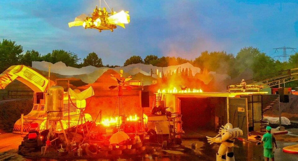 scene foto robot Een witte robot en een vrouw gekleed in groen, aanschouwen de werkplaats die afbrand, bovenin vliegt een voertuig met mensen.