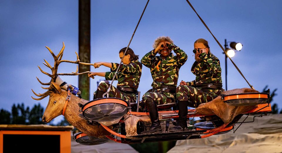 Drie mensen zitten in camouflage kleding op een vliegend voertuig met een grote hertenkop en gewei als stuur. De middelste persoon houdt diens handen als een verrekijker.