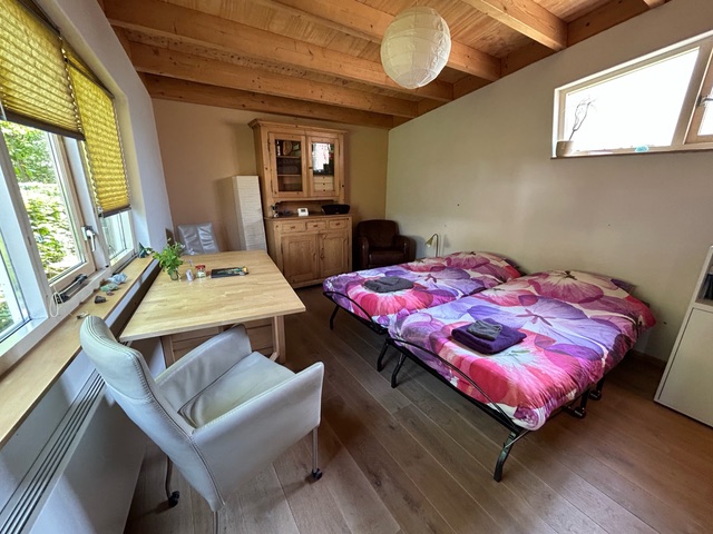 Zicht op de binnenkant van een huisje, er staat een houtkleurige kast tegen de muur achterin.  Verder staan er twee rozige opgemaakte bedden, een bureau en een witte stoel.
