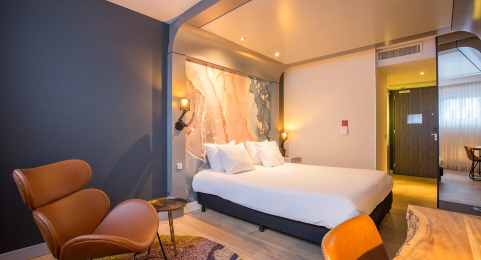 Foto van een luxe kamer met een tweepersoonsbed tegen een muur. Verder staat er een bruine stoel en een houten bureau.